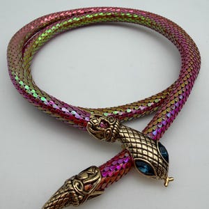 Puede incluir: Un collar flexible con forma de serpiente que presenta un cuerpo de malla tubular con un acabado iridiscente en rosa, morado y verde. La pieza incluye dos cabezas de serpiente de metal en tono dorado antiguo con ojos de gema azul y lengua bífida.