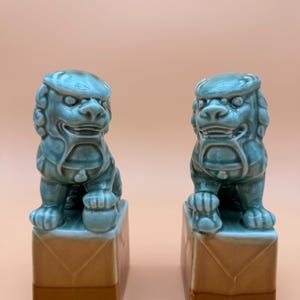 May include: Two turquoise ceramic Foo Dog statues. Each dog is seated atop a brown square base. The Foo Dogs have detailed features, including curly manes and expressive faces. The background is a solid, light peach color.