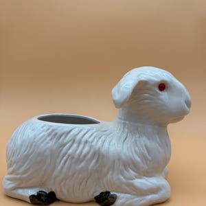 Peut inclure: Un pot de fleurs en céramique blanche en forme d'agneau, avec des yeux rouges et des sabots noirs. L'agneau est en position de repos, avec une grande ouverture sur le dos pour planter. Le pot a un aspect texturé, semblable à de la fourrure.