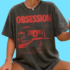 May include: A charcoal gray short-sleeve t-shirt featuring the word OBSESSION in large red letters. Below the text is a red graphic of a house with a figure in the doorway and a car in the foreground, followed by a dictionary definition of obsession.