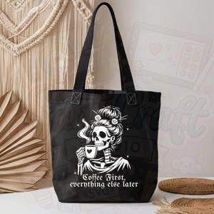 May include: Black tote bag featuring a white graphic of a skeleton woman sipping coffee, with the text "Coffee First, everything else later." The bag has black handles and is likely made of canvas or a similar material.