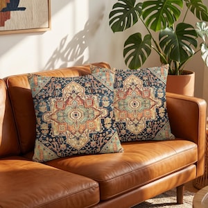 Puede incluir: Dos cojines decorativos cuadrados con un patrón geométrico azul marino, óxido y beige. Los cojines, con un diseño que recuerda a una alfombra tradicional, están colocados sobre un sofá de cuero marrón. El patrón añade un toque de elegancia al salón.