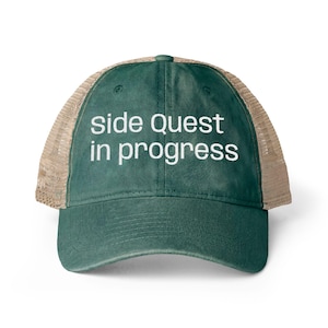 Può includere: Un cappellino da camionista verde con retro in rete beige. La parte anteriore del cappello presenta il testo bianco "side quest in progress". Il cappello ha una visiera curva e un bottone sulla parte superiore.