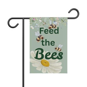 Puede incluir: Una bandera de jardín con un fondo verde claro, con el texto "Feed the Bees" en verde oscuro. La bandera tiene ilustraciones de abejas y una gran margarita. La bandera está sujeta a una estaca de jardín de metal negro.