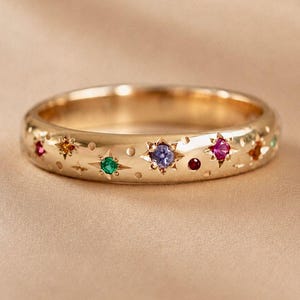 May include: A gold band ring featuring a series of flush-set multicolored gemstones, including green, purple, red, and orange stones. The metal surface is engraved with starburst patterns surrounding each stone, creating a celestial design.