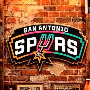 May include: Wooden San Antonio Spurs sign with the team logo in white, black, orange, pink, and teal. The sign is mounted on a brick wall. The sign reads "SAN ANTONIO SPURS".