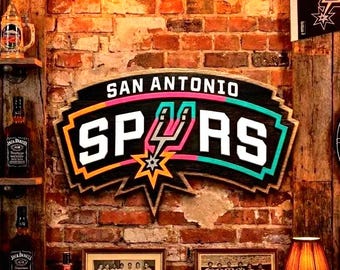 May include: Wooden San Antonio Spurs sign with the team logo in white, black, orange, pink, and teal. The sign is mounted on a brick wall. The sign reads "SAN ANTONIO SPURS".