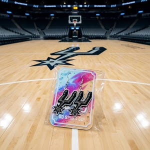 Op de afbeelding: Een paar zwarte en zilveren oorbellen in de vorm van het logo van de San Antonio Spurs, gepresenteerd op een kleurrijke kaart met de tekst "GO SPURS GO!". De oorbellen zitten in een doorzichtige plastic verpakking, tegen een achtergrond van een basketbalveld.