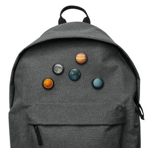 Könnte beinhalten: Ein grauer Rucksack mit mehreren Anstecknadeln zum Thema Planeten. Die Anstecknadeln zeigen Sonne, Mond, Erde, Jupiter und Uranus. Der Rucksack hat eine Vordertasche und einen schwarzen Griff.