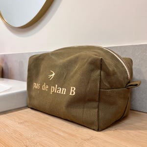 Op de afbeelding: Olijfgroene canvas toilettas met een witte rits en de tekst "pas de plan B" in goud. Een kleine gouden vogelafbeelding staat boven de tekst. De rechthoekige tas ligt op een houten oppervlak.
