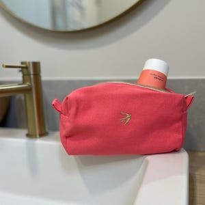 Op de afbeelding: Een koraalkleurige canvas make-up tas met een gouden rits en een klein, goud geborduurd vogel embleem. Een tube product is gedeeltelijk zichtbaar. De tas staat op een witte wastafel, met een gouden kraan op de achtergrond.