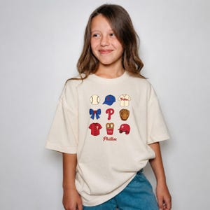 May include: A cream-colored short-sleeve t-shirt featuring a graphic print of baseball-themed icons including a ball, cap, jersey, bow, the letter P, glove, pretzel, and helmet, with the word Phillies printed in red script below the icons.
