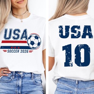 K&ouml;nnte beinhalten: Wei&szlig;es T-Shirt mit "USA SOCCER 2026" Grafik. Das Design auf der Vorderseite zeigt das Wort "USA" in Blau, einen Fu&szlig;ball und rot-blaue Streifen. Auf der R&uuml;ckseite des Shirts stehen "USA" und die Nummer "10" in Blau.