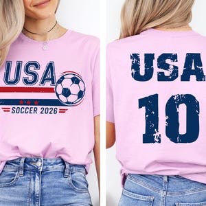 K&ouml;nnte beinhalten: Hellrosa T-Shirt mit dem Text "USA" in Marineblau, einem rot-blauen Streifendesign, einer Fu&szlig;ballgrafik und dem Text "SOCCER 2026". Auf der R&uuml;ckseite des Shirts stehen "USA" und die Nummer "10" in Marineblau.