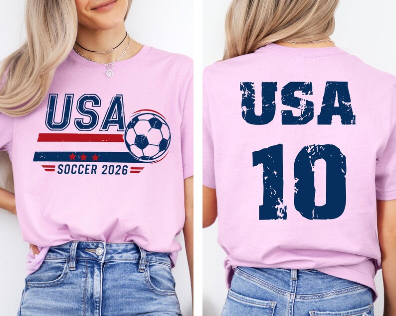 K&ouml;nnte beinhalten: Hellrosa T-Shirt mit dem Text "USA" in Marineblau, einem rot-blauen Streifendesign, einer Fu&szlig;ballgrafik und dem Text "SOCCER 2026". Auf der R&uuml;ckseite des Shirts stehen "USA" und die Nummer "10" in Marineblau.