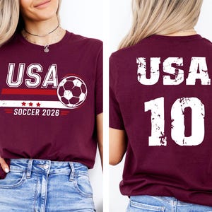 K&ouml;nnte beinhalten: Burgunderfarbenes T-Shirt mit "USA" in Wei&szlig;, einer Fu&szlig;ballgrafik und dem Text "SOCCER 2026". Auf der R&uuml;ckseite des Shirts stehen "USA" und die Nummer "10" in Wei&szlig;.