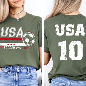 K&ouml;nnte beinhalten: Olivgr&uuml;nes T-Shirt mit "USA" in wei&szlig;en Blockbuchstaben, einer Fu&szlig;ballgrafik und dem Text "SOCCER 2026". Auf der R&uuml;ckseite des Shirts stehen "USA" und die Nummer "10" in Wei&szlig;. Das Shirt besteht aus einem weichen Material.