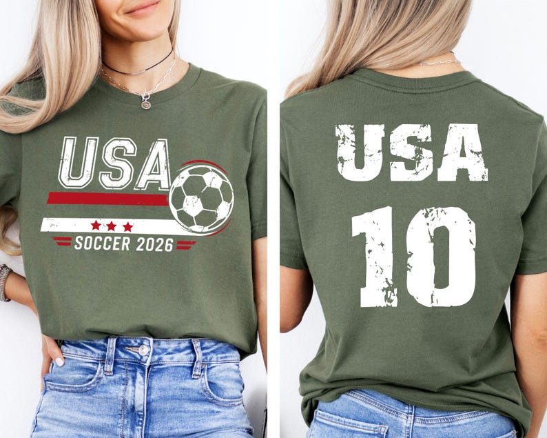 K&ouml;nnte beinhalten: Olivgr&uuml;nes T-Shirt mit "USA" in wei&szlig;en Blockbuchstaben, einer Fu&szlig;ballgrafik und dem Text "SOCCER 2026". Auf der R&uuml;ckseite des Shirts stehen "USA" und die Nummer "10" in Wei&szlig;. Das Shirt besteht aus einem weichen Material.