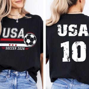 K&ouml;nnte beinhalten: Schwarzes T-Shirt mit "USA" in wei&szlig;en Buchstaben, einer Fu&szlig;ballgrafik und "SOCCER 2026" in Rot und Wei&szlig;. Auf der R&uuml;ckseite des Shirts stehen "USA" und die Nummer "10" in Wei&szlig;.