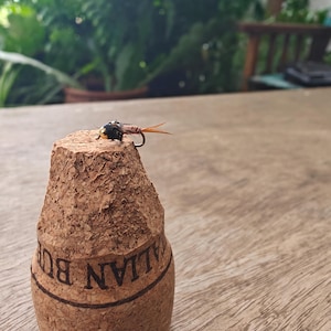 Könnte beinhalten: Nahaufnahme eines Korken mit einer Angel-Fliege. Der Korken ist braun mit einer strukturierten Oberfläche und dem Aufdruck "Italian Burgundy" an der Seite. Die Fliege ist schwarz, braun und orange.