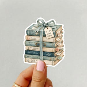 Puede incluir: Una pegatina que muestra una pila de seis libros en tonos azules atados con una cinta azul claro y una pequeña etiqueta rectangular que dice TO BE READ. Los libros tienen lomos con texto visible y pequeños marcapáginas laterales.