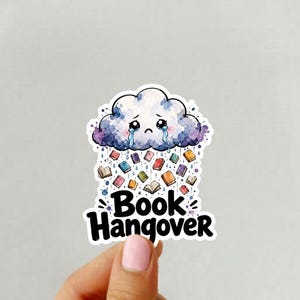 May include: A die-cut sticker featuring a sad, crying purple and blue cloud raining small colorful books. Below the cloud, the text reads Book Hangover in bold black letters. The design uses a watercolor style with a white border around the edges.