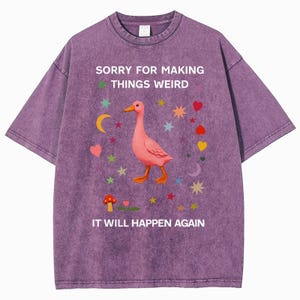 May include: Purple t-shirt with the text "SORRY FOR MAKING THINGS WEIRD IT WILL HAPPEN AGAIN." The shirt features a pink duck illustration surrounded by colorful stars, hearts, and a mushroom.