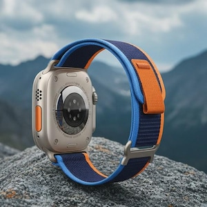 May include: A silver smartwatch with an orange and blue band rests on a gray rock. The watch has a circular screen and a series of buttons on the side. The band is made of a woven material and has a metal clasp. Mountains and a cloudy sky are in the background.
