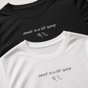 Op de afbeelding: Een zwart t-shirt en een wit t-shirt, beide voorzien van de tekst "sweet 'n a lil' spicy" in een cursief lettertype, vergezeld van een kleine lijntekening van een lolly en een chilipeper gecentreerd onder de tekst.