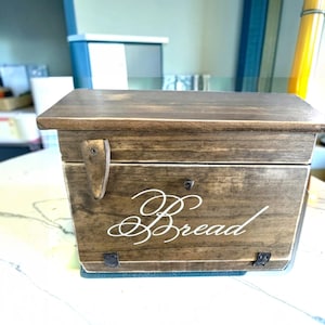 May include: A rectangular wooden bread box with a dark brown stained finish and a hinged lid. The front panel features the word Bread in white cursive script, a small metal latch on the left side, and two small metal hinges at the bottom edge.