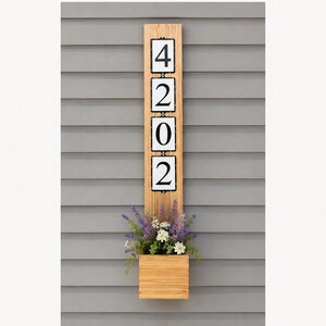 May include: A vertical light brown wooden plaque featuring four white square tiles with black borders and the black numbers 4, 2, 0, and 2 stacked vertically. A small wooden planter box is attached at the bottom, containing artificial purple and white flowers.