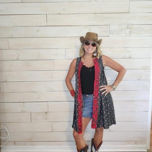 May include: A sleeveless charcoal gray duster vest featuring a white star pattern and bright red lace trim along the front opening. The garment is worn over a black tank top and a blue denim skirt, paired with brown leather cowboy boots and a hat.