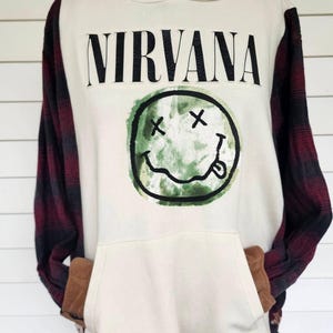 May include: A cream-colored hooded sweatshirt featuring the word NIRVANA in black text above a green watercolor-style smiley face graphic. The garment has long sleeves made of a red and black plaid flannel fabric and brown pocket trim.