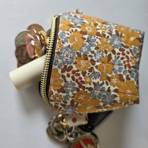 Könnte beinhalten: Ein kleines, gestepptes Stofftäschchen mit einem Blumenmuster in Senfgelb, Hellblau und Weiß. Das Täschchen hat einen goldfarbenen Reißverschluss und enthält mehrere Metallmünzen, einen rosafarbenen blumenförmigen Anhänger und eine weiße Tube.