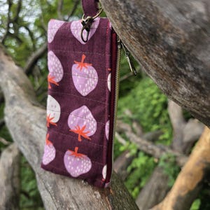 May include: A burgundy fabric wallet with a strawberry pattern, featuring light purple strawberries with orange stems and white seeds. The wallet has a zipper closure and a small strap with a metal clasp. The background is a blurred view of a tree and foliage.