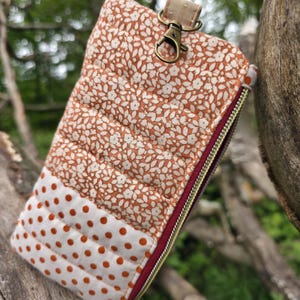 May include: A quilted fabric pouch with a floral pattern in shades of orange and white, and a section of white fabric with orange polka dots. It has a burgundy zipper and a gold-colored clasp and strap.