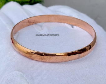 May include: A polished copper bangle bracelet with a smooth, rounded surface. The metal has a warm, reddish-brown metallic lustre. The text SILVERLEGANCEJAIPUR is printed in black on the surface of the bangle, which is resting on a white fabric.