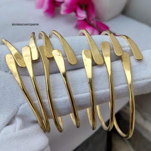 Pode incluir: Um conjunto de pulseiras de metal dourado com acabamento liso e polido e extremidades afuniladas. As pulseiras apresentam um design aberto e estão dispostas em fila. O texto SILVERLEGANCEJAIPUR é visível no canto superior esquerdo.