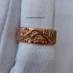 Op de afbeelding: Een koperkleurige metalen ring met een reliëf bloemenpatroon van krullende ranken en kleine bloemetjes. De band heeft een textuurafwerking en de tekst SILVERLEGANCEJAIPUR is zichtbaar boven de ring op de witte stoffen achtergrond.