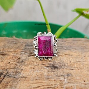Può includere: Un anello con una pietra preziosa rettangolare rosa scuro incastonata in una cornice decorativa in metallo argentato con piccoli accenti circolari. La montatura in metallo presenta un bordo smerlato attorno alla pietra. Il testo SILVERLEGANCEJAIPUR è visibile.