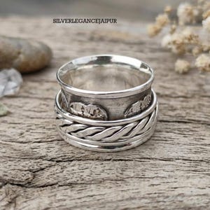 Peut inclure: Une bague rotative en argent avec une bande centrale ornée de silhouettes de montagnes en relief et une bande inférieure tressée. La bague présente une finition polie avec des zones en creux assombries. Le texte indique SILVERLEGANCEJAIPUR.