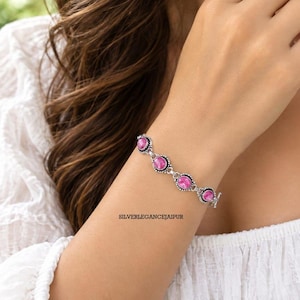 Peut inclure: Un bracelet en chaîne argentée orné de quatre pierres précieuses ovales d'un rose vif, serties dans des montures métalliques décoratives. Le bracelet comporte un fermoir à bascule et le texte SILVERELEGANCEJAIPUR est visible près du bijou.