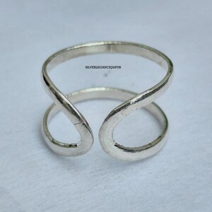 May include: A silver-coloured open-band ring featuring a smooth, polished finish and a continuous, curved design that creates a gap at the front. The text SILVERLEGANCEJAIPUR is printed in small black letters on the upper portion of the metal band.