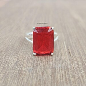 May include: A silver ring featuring a large, rectangular, faceted red gemstone held by four prongs. The band is smooth and metallic. The text SILVERLEGANCEJAIPUR is printed in small black letters above the center of the red gemstone.