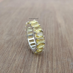 Op de afbeelding: Een zilveren eternity-ring met een doorlopende rij rechthoekige, smaragdgeslepen gele edelstenen in een klauwzetting. De ring bevat de tekst SILVERELEGANCEJAIPUR aan de binnenzijde. Het ontwerp toont een zilverkleurige afwerking met gele stenen.