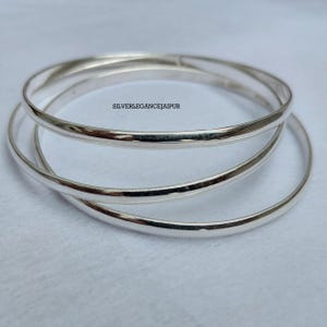 May include: A set of three interlocking silver bangles with a smooth, polished finish. The text SILVERLEGANCEJAIPUR is visible on the surface of the top bangle. These circular bracelets feature a simple, thin design with a reflective metallic surface.