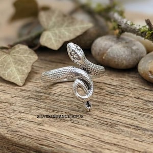 Può includere: Un anello in metallo color argento a forma di serpente avvolto con squame testurizzate che coprono l'intera superficie. La testa del serpente è posizionata in alto, mentre la coda si curva verso l'interno per formare l'anello. Il testo SILVERLEGANCEJAIPUR è visibile.