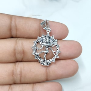 Könnte beinhalten: Ein silberner Anhänger mit einer detaillierten Darstellung der hinduistischen Gottheit Shiva in der Nataraja-Pose, umgeben von einem dekorativen Ring mit flammenartigen Motiven. Der Anhänger hat eine Öse und den Text SILVERELEGANCEJAIPUR.