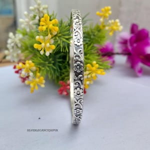 Könnte beinhalten: Ein silberner Armreif mit einem durchgehenden Prägemuster aus kleinen Blumen und geschwungenen Ranken. Die Metalloberfläche weist in den vertieften Bereichen ein dunkleres Finish auf, um die floralen Details hervorzuheben. Der Text SILVERLEGANCEJAIPUR ist unten sichtbar.