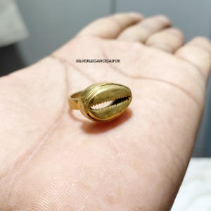 Op de afbeelding: Een goudkleurige ring met een kaurischelp-ontwerp op de voorzijde. De metalen band is glad en cirkelvormig. De tekst SILVERLEGANCEJAIPUR is zichtbaar op de achtergrond boven de ring, die op een handpalm rust.